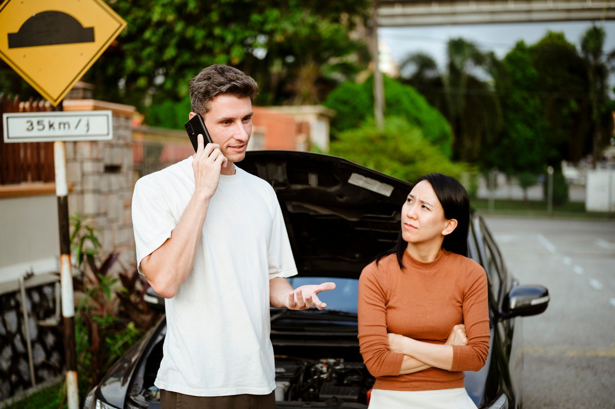 Husband calling for roadside assistance during car trouble. Driver speaking on the phone beside a disabled rental car after an accident in Illinois while a passenger looks concerned.